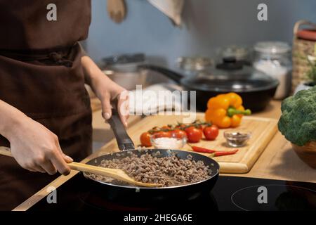 Hände einer Frau in der Schürze, die einen Holzlöffel hielt, während sie am Elektroherd stand und Hackfleisch in einer Pfanne mischte Stockfoto