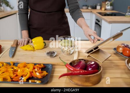 Hand einer jungen Frau in der Schürze, die auf den Tablet-Bildschirm zeigt, während sie am hölzernen Küchentisch steht und frisches Gemüse hackt Stockfoto