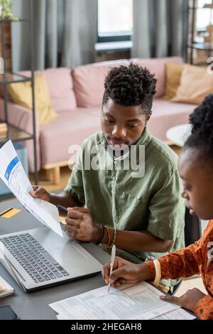 Junger afrikanischer Ökonom mit Finanzpapier und Stift, der am Arbeitsplatz mit einer Kollegin interagiert Stockfoto