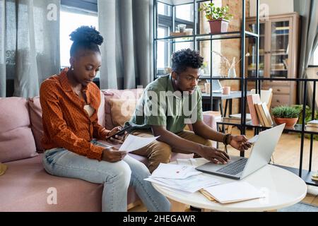 Junge schwarze Frau, die auf dem Sofa sitzt und durch die Finanzzeitung schaut, während ein afrikanischer Mann auf das Notebook-Display schaut Stockfoto