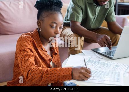 Junge ernsthafte afrikanische Frau, die Notizen in Finanzdokumenten macht, während sie gegen einen Mann sitzt, der vor dem Laptop netzt Stockfoto