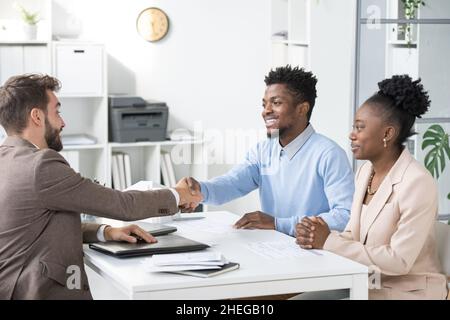 Junger eleganter Anwalt in formalwear gratuliert dem afrikanischen Paar, während er einem glücklichen Mann die Hand über den Tisch im Büro schüttelt Stockfoto