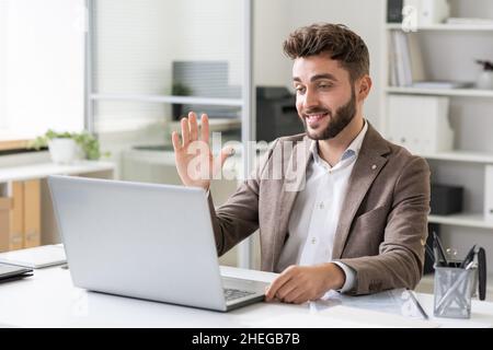 Glücklicher junger Geschäftsmann, der jemandem auf dem Laptop-Bildschirm die Hand winkt, während er während eines Online-Meetings kommuniziert Stockfoto