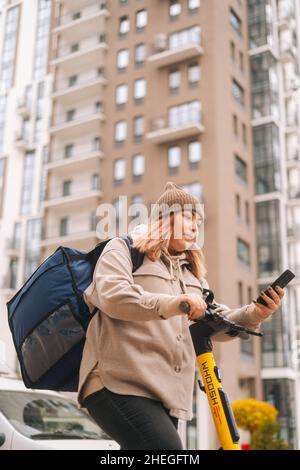 Vertikale Aufnahme der Lieferung Frau, die auf dem Handy in der Nähe von Elektroroller auf städtischen Spielplatz. Stockfoto