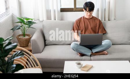 Technologiekonzept der Mann, der in legerer Kleidung auf dem Sofa sitzt, sein Notebook auf dem Schoß auslaht und seine Arbeit verrichten will. Stockfoto