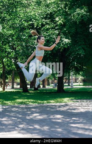 Lächelnde junge, sportliche Frau, die in der Luft springt und die Arme freudig in grauer Sportkleidung ausstreckt, am sonnigen Sommertag im Park Stockfoto