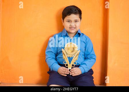 Portrait of happy adorable niedlichen indischen Jungen in der Schuluniform feiert Sieg mit Trophäe, fröhlich männlichen Kind hält Gewinnpreis gegen orange BA Stockfoto