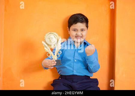 Glücklich liebenswert niedlich indischen Jungen in der Schuluniform feiert Sieg mit Trophäe, fröhlich aufgeregt männlichen Kind hält Gewinnpreis gegen orange backgr Stockfoto