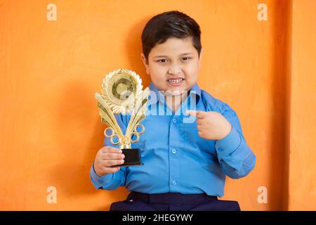 Portrait of happy adorable niedlichen indischen Jungen in der Schuluniform feiert Sieg mit Trophäe, fröhlich männlichen Kind hält Gewinnpreis gegen orange BA Stockfoto