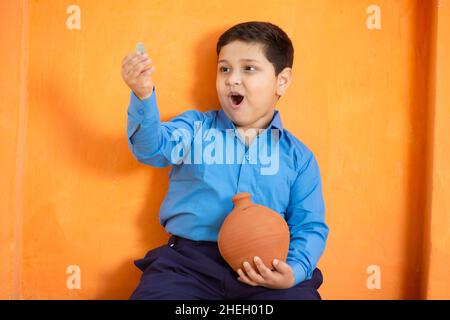 Happy niedlichen kleinen indischen Jungen mit Münze in der Hand und Ton-Geld-Box oder gullak in der anderen Hand.Überraschte männliche Kind mit traditionellen Sparschwein, Schule ch Stockfoto