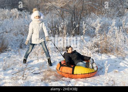 Die ältere Schwester und ihr kleiner Bruder reiten auf dem Schneeröhr am sonnigen Wintermorgen. Glückliche Kinder Gesichter. Lustige Spiele im Winterurlaub. Alles ist weiß, f Stockfoto