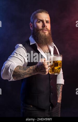 Porträt eines Bartmenschen, der einen Becher Bier hält und die Kamera auf schwarzem Hintergrund anschaut Stockfoto