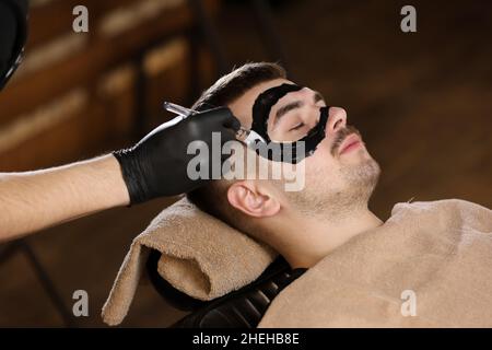 Anwendung einer schwarzen Maske auf das Gesicht eines Mannes. Mann in Einweghandschuhen trägt eine schwarze Maske auf das Gesicht des Mannes. Hautpflege. Stockfoto