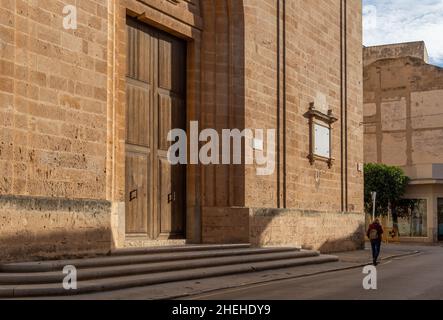 Hauptfassade der Pfarrkirche von Sant Miquel, in der mallorquinischen Stadt Llucmajor, im Morgengrauen und mit einer unkenntlichen Person, die die Straße entlang läuft Stockfoto