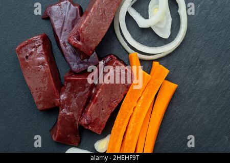 Stücke rohes Leberfleisch mit Karotte und Zwiebel auf der Steinplatte, Draufsicht, isoliert auf Weiß Stockfoto