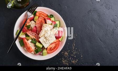 Rettich, Gurke, Tomaten, Pfeffer und Feta-Käse mit Gewürzen Pfeffer und Olivenöl in einer weißen Schüssel auf schwarzem Schiefer, Stein oder Beton. Gesund Stockfoto