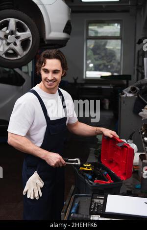 Lächelnder Mechaniker, der in der Nähe von Werkzeugen und Werkzeugkiste im Autoservice steht Stockfoto