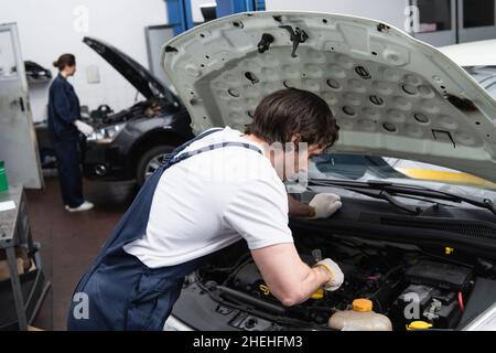 Vorarbeiter in Handschuhen und Overalls, die mit einem Auto mit offener Motorhaube in der Garage arbeiten Stockfoto