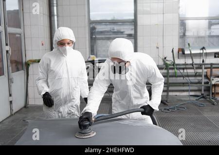 Arbeiter in Hazmat Anzug und Atemschutzmaske Polieren Auto in der Nähe Kollegen in der Garage Stockfoto