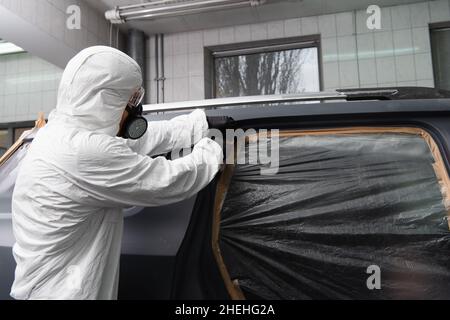 Arbeiter in Atemschutzmaske und Gefahrgutanzug, der Klebeband auf Zellophan auf dem Auto im Betrieb aufklebt Stockfoto