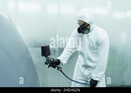 Arbeiter in Gefahrgut-Anzug und Atemschutzmaske mit Aerograph auf Autoteil in der Garage Stockfoto