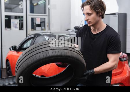 Junger Arbeiter in Handschuhen, der Reifen in der Nähe von verschwommenem Auto hält, das in Betrieb ist Stockfoto