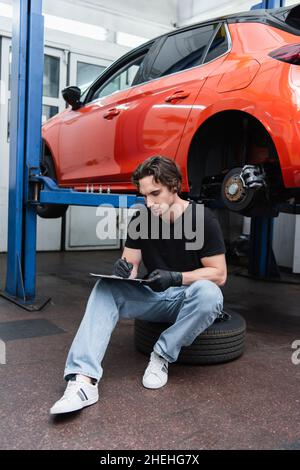 Mechaniker in Handschuhen, die auf der Zwischenablage schreiben, während er in der Nähe des Autos in der Garage auf dem Reifen sitzt Stockfoto