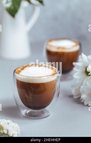 Zwei Tassen Kaffee in einem Glas Tassen auf einem grauen Holzhintergrund mit Blumendekoration. Caffe Latte oder Espresso Macchiato. Stockfoto
