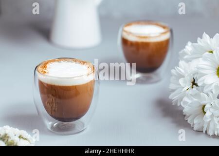 Zwei Tassen Kaffee in einem Glas Tassen auf einem grauen Holzhintergrund mit Blumendekoration. Caffe Latte oder Espresso Macchiato. Stockfoto