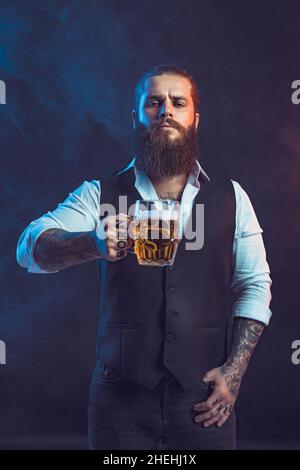 Porträt eines Bartmenschen, der einen Becher Bier hält und die Kamera über dem Rauchschwaden anschaut. Tätowierungen Hände Stockfoto