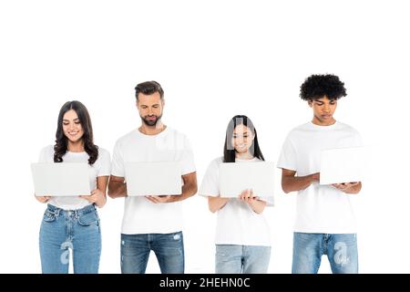 Lächelnde, multiethnische Freiberufler in T-Shirts und Jeans mit Laptops, die auf Weiß isoliert sind Stockfoto