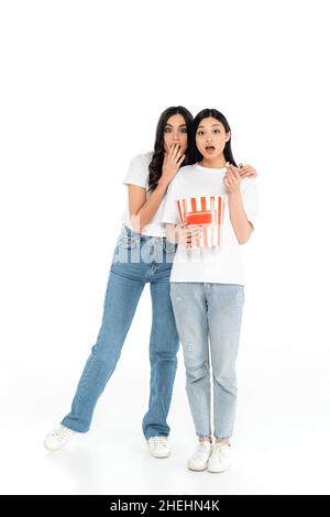 Schockierte Frau, die den Mund mit der Hand bedeckte, während sie einen Film mit einem asiatischen Freund sah, der einen Eimer Popcorn auf Weiß hielt Stockfoto
