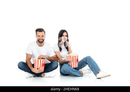 In voller Länge sehen Sie ein glückliches Paar in Jeans, das Popcorn isst, während es auf Weiß sitzt Stockfoto