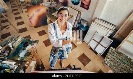 Fröhliche Künstlerin lächelt in ihrem Atelier. Fantasievolle junge Malerin, die mit gekreuzten Armen auf die Kamera schaut. Kreative junge wo Stockfoto