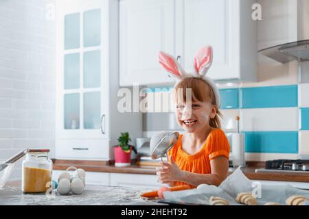 Emotionales Porträt eines fröhlichen kleinen Mädchens, das zu Ostern als Hase gekleidet ist, während man zu Hause in der Küche Essen kocht. Mädchen Kind Kind mit Spaß lacht spielt Stockfoto