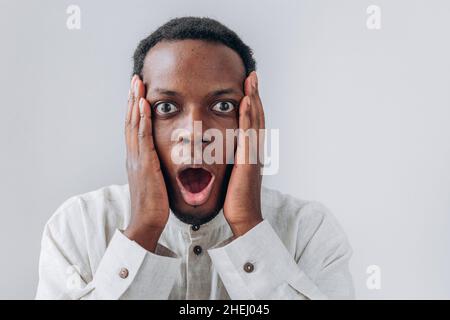 Porträt eines lustig schockierten afroamerikanischen Mannes in weißem Hemd mit Wangen, die auf hellgrauem Hintergrund stehen, Nahaufnahme Stockfoto