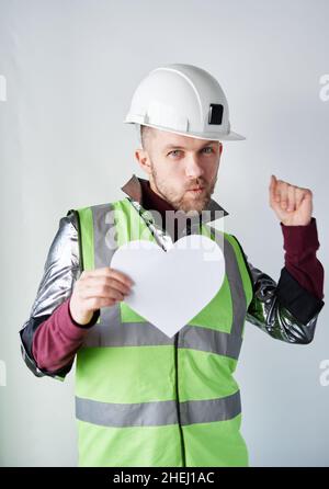 Valentinstag: tanzender bärtiger männlicher Bauarbeiter oder Techniker in einem Hut und einer reflektierenden Weste, die eine Herzform aus weißem Papier hält. Romantik oder Beziehungskonzept. Hochwertige Bilder Stockfoto