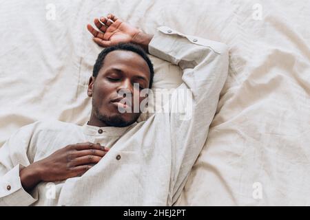 Schöner ruhiger afroamerikanischer Mann in weißem Leinenhemd schläft auf einem bequemen Bett in hellem Raum zu Hause aus der oberen Ansicht Stockfoto