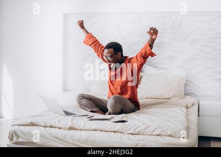 Fröhlicher afroamerikanischer Kerl in legerer Kleidung und Brille legt die Hände hoch, die in der Nähe eines offenen Laptops auf einem bequemen Bett zu Hause sitzen Stockfoto