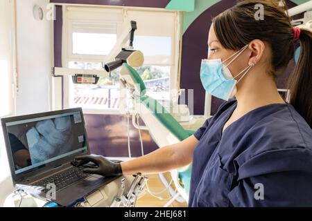 Die junge Ärztin, die eine Maske trägt, sieht sich in ihrer Zahnarztpraxis eine Röntgenaufnahme am Computer an Stockfoto