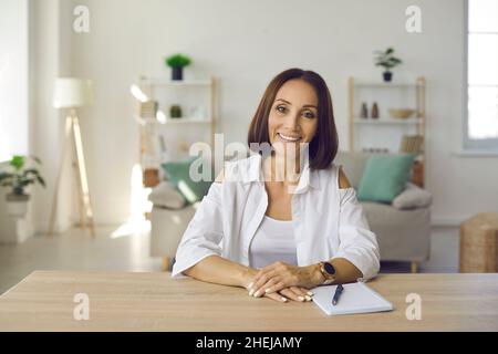 Fröhliche lächelnde Frau mittleren Alters im lässigen Hemd, die zu Hause mit einem Notizbuch am Schreibtisch sitzt Stockfoto