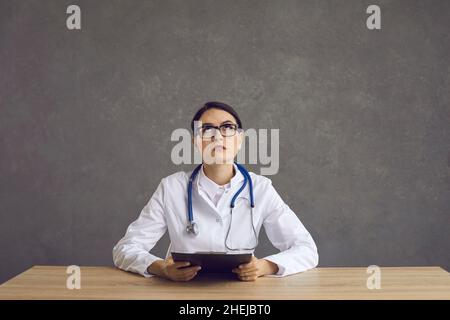 Porträt einer ernsthaften jungen Ärztin, die an einem Schreibtisch sitzt, aufschaut und nachdenkt Stockfoto