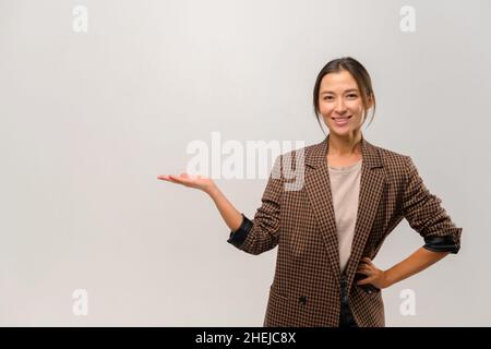 Lächelnde multirassische Geschäftsfrau präsentiert Neuheit isoliert auf grauem Hintergrund, charmante asiatische Frau in stilvollen lässig gelötet halten beworbenen Produkt auf der Handfläche, Kopierraum Stockfoto