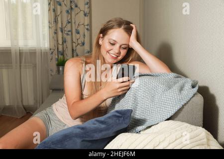 Glückliche junge blonde Frau mit Smartphone, während sie zu Hause auf einer Couch sitzt. Stockfoto