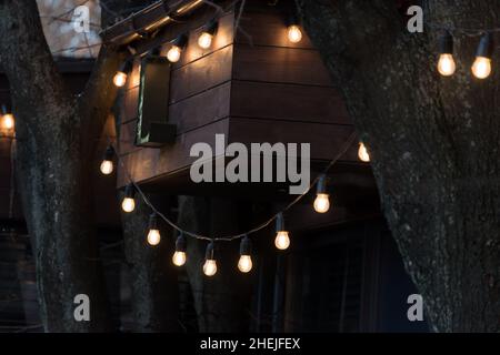 Verschwommener Hintergrund, Hinterhofbeleuchtung, Licht im Abendgarten, elektrische Laternen mit rundem Diffusor. Lampe Girlande von Glühbirnen, beleuchten Stockfoto