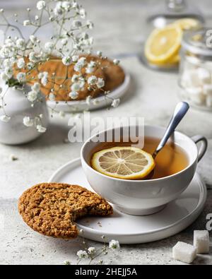 Weiße Porzellanbecher mit Zitronentee und Haferflocken-Cookies auf strukturiertem Hintergrund mit Blumen auf der Rückseite Stockfoto