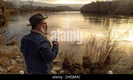 Der Wanderer genießt eine wunderschöne natürliche Landschaft mit einer Tasse heißem Getränk. Mann trinkt Kaffee und bewundert den wunderschönen Fluss bei Sonnenaufgang. Stockfoto