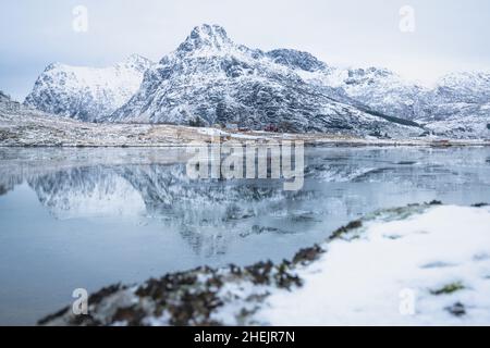 Gefrorener Fjord mit schneebedeckten Bergen, die sich im Wasser spiegeln, Flakstadpollen, Flakstadoya, Nordland, Lofoten-Inseln, Norwegen Stockfoto