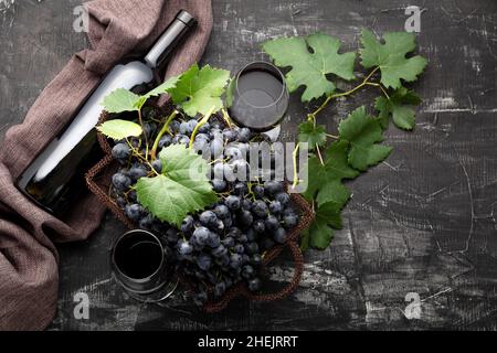 Rotwein-Flasche und Korb mit schwarzen Trauben und ein Glas Rotwein auf dunklem rustikalem Beton-Hintergrund. Flaches Lay Weinzusammensetzung Rotwein Flasche auf Stockfoto