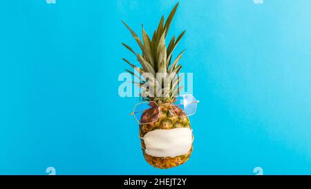 Ananas lustiges Gesicht in Sonnenbrille medizinische Maske auf Farbe blau Sommer Hintergrund. Reisekonzept Ananas in Schutzmaske in Coronavirus-Lockdown Stockfoto